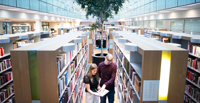 Två studenter som tittar i en bok på ett bibliotek. 